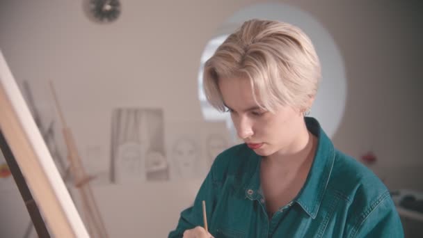 Uma jovem loira com artista de cabelo curto desenha uma pintura no estúdio de arte — Vídeo de Stock