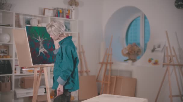 En ung kvinnelig kunstner legger til ferdig berører maleriet og setter seg på stolen – stockvideo