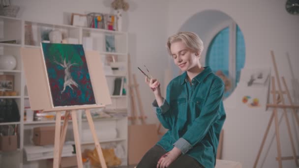 En ung kvinnelig kunstner sitter på stolen i et kunststudio med pensler – stockvideo