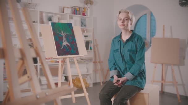 Молода жінка-художниця з коротким світлим волоссям сидить у художній студії після закінчення картини — стокове відео