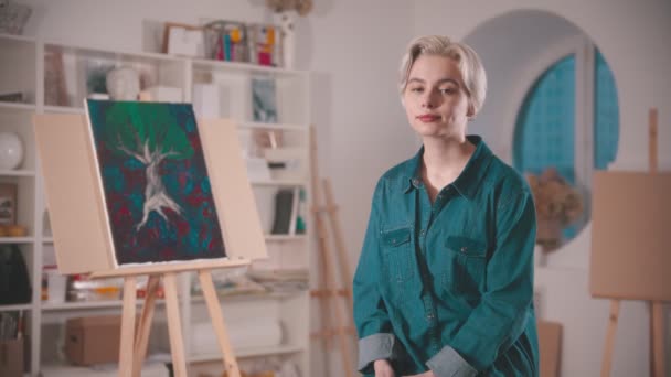 Een jonge vrouw artiest met kort blond haar zittend in de kunststudio — Stockvideo