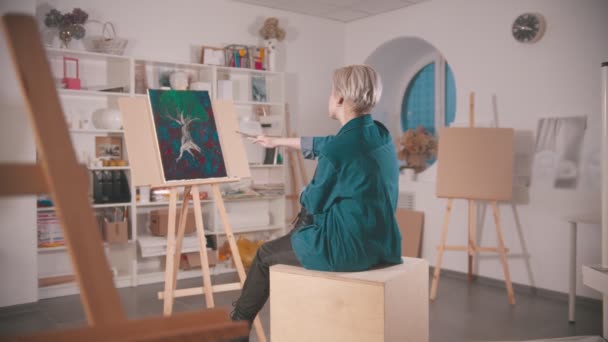 Una joven con el pelo corto y rubio sentada en el estudio de arte mirando su pintura desde la distancia sosteniendo pinceles — Vídeo de stock