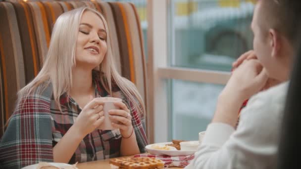 Egy fiatal szőke nő kávét iszik és a barátjával beszél a kávézóban. — Stock videók