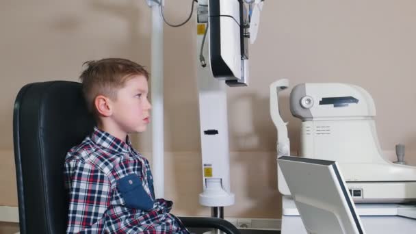 Лечение офтальмологом - маленький мальчик проверяет глаза с большим устройством за столом — стоковое видео