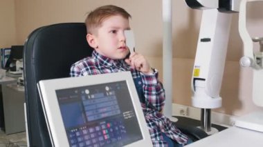 Klinikte oftalmolog tedavisi gören küçük bir çocuk gözünü bir göz kalkanıyla kapatıyor ve masadan gelen mektupları okuyor.