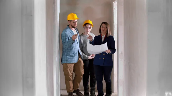 Un agent immobilier montrant un nouveau projet d'appartement à un jeune couple marié dans des casques - regarder autour — Photo