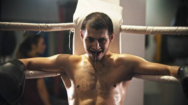 Ağzında diş koruyucusuyla ringde oturan yorgun, gülümseyen bir boksör.