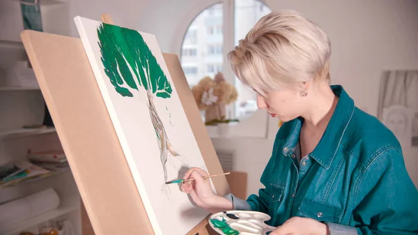 A young blonde woman draws a tree using colorful paints — 스톡 사진