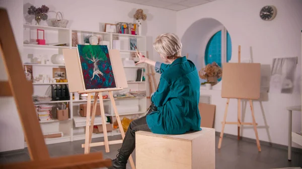 Μια νεαρή γυναίκα με κοντά ξανθά μαλλιά κάθεται στο στούντιο τέχνης - κοιτάζοντας τη ζωγραφική της από απόσταση κρατώντας βούρτσες — Φωτογραφία Αρχείου