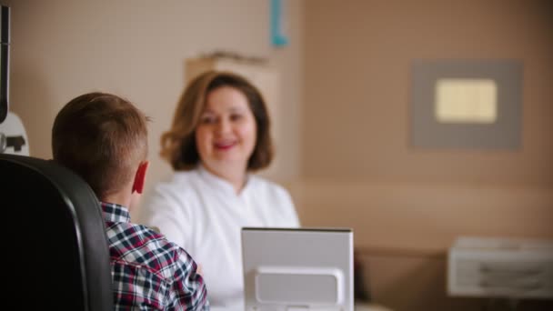 Ophthalmologist treatment - mature woman doctor sitting by the table with a little boy - giving the boy an eye shield — Stock Video