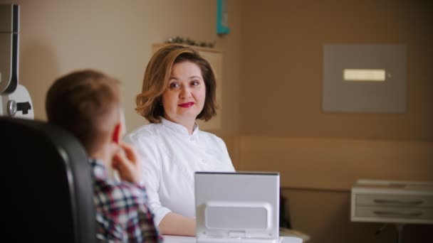 Лечение офтальмологом - зрелая женщина-врач сидит за столом с маленьким мальчиком, смотрящим на проекцию стен — стоковое видео