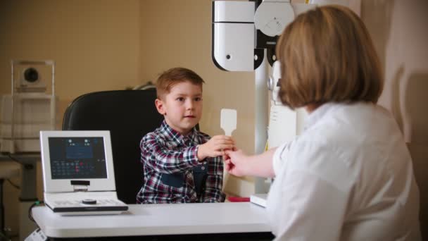 Um tratamento na clínica ocular mulher madura médico sentado à mesa com um menino dando ao menino um escudo ocular — Vídeo de Stock