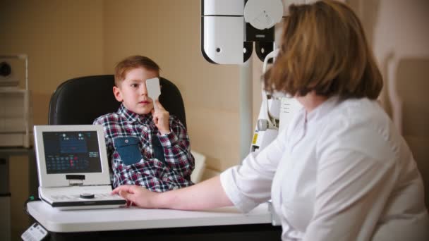 Een behandeling in oogkliniek - een kleine jongen die zijn oog bedekt met een oogschild en brieven leest van de projectie op de muur — Stockvideo