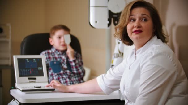 Un traitement à la clinique ophtalmologique - un petit garçon qui se couvre l "œil avec un bouclier oculaire et lit les lettres de la projection sur le mur - une femme médecin qui le suit — Video