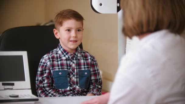 Μια θεραπεία στην οφθαλμολογική κλινική - ώριμη γυναίκα γιατρός κάθεται στο τραπέζι με ένα μικρό αγόρι — Αρχείο Βίντεο