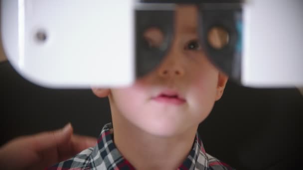 Een behandeling in oogkliniek - het controleren van kleine jongens oog zicht door te kijken door middel van grote speciale apparaat met verwisselbare lenzen — Stockvideo