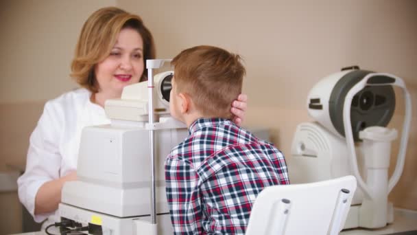 El niño que recibe un tratamiento en la clínica oftalmológica: una doctora que revisa la visión ocular de los niños pequeños mirando a través de un dispositivo especial grande — Vídeos de Stock