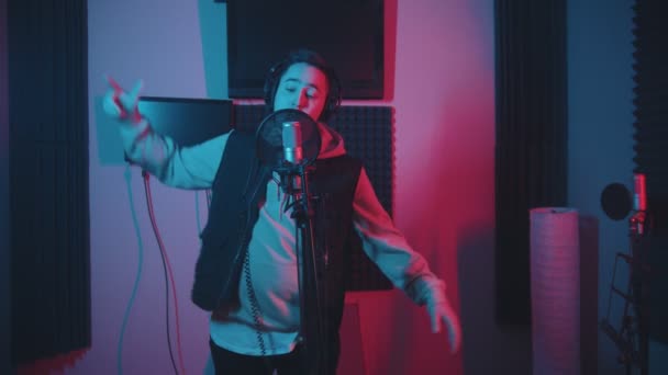 Egy kapucnis férfi rappel a stúdióban gesztikulál, miközben felveszi a zenéjét. — Stock videók