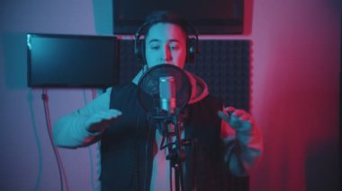Plak stüdyosunda rap yapan kapüşonlu bir adam şarkısını kaydederken el kol hareketi yapıyor.
