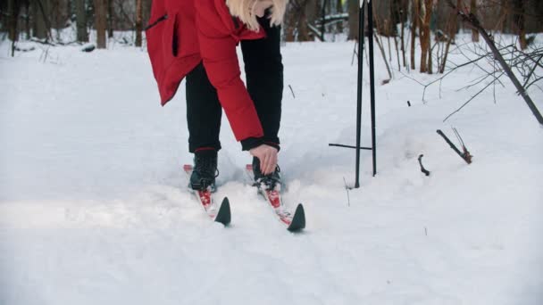A young blonde woman in red down jacket unsetting her boots from the ski — ストック動画