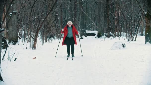 Een jonge blonde vrouw in rood donsjas wandelen op ski in het bos — Stockvideo