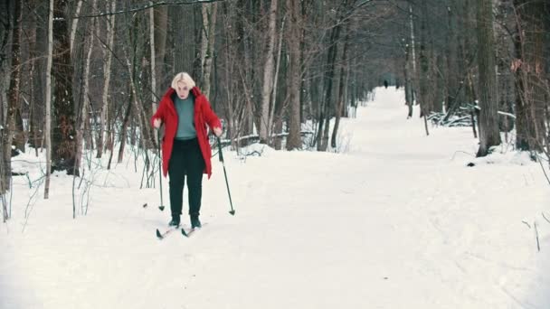 A young blonde woman in red down jacket skiing in the forest — ストック動画