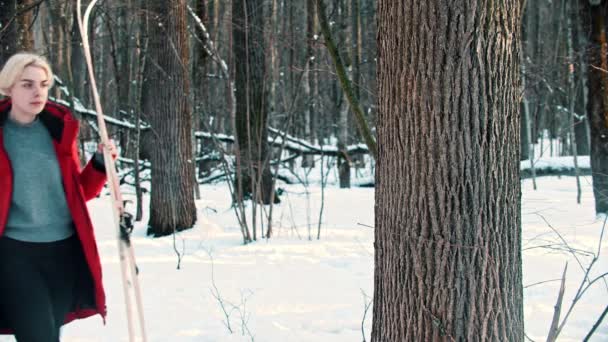 Молодая блондинка в красной куртке положила лыжи возле дерева в лесу. — стоковое видео