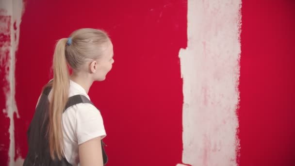 Жінка в комбінезоні малює червону стіну з білим валиком — стокове відео