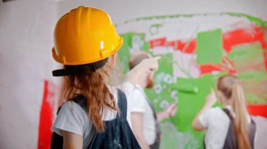 Girl in overalls and a yellow helmet is telling parents where to paint the wall