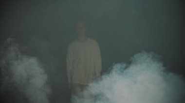 Young man dancer walking out from the smoke in the dark studio