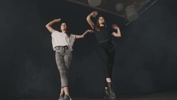 Две молодые привлекательные женщины тренируют синхронную танцевальную хореографию в темной студии — стоковое видео