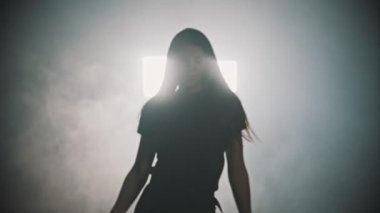A young attractive woman with long hair walking out from the smoke and start dancing on a background of bright lighting