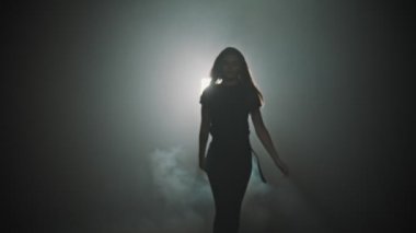 A young attractive woman walking out from the smoke and start dancing on a background of bright lighting