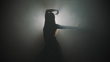 A silhouette of young attractive woman dancing with her hands on the background of bright lighting