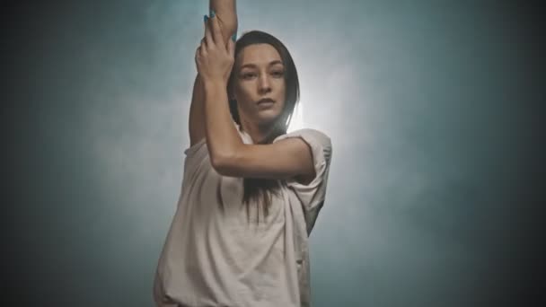 Une femme flexible en t-shirt blanc dansant avec ses mains dans le studio fumé — Video