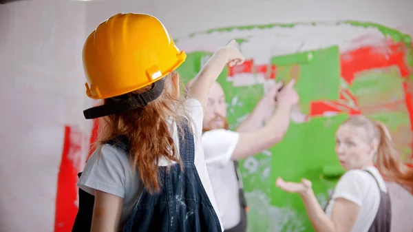 Een klein meisje in een helm wijst naar de muur - haar verbaasde ouders staan naast de muur — Stockfoto