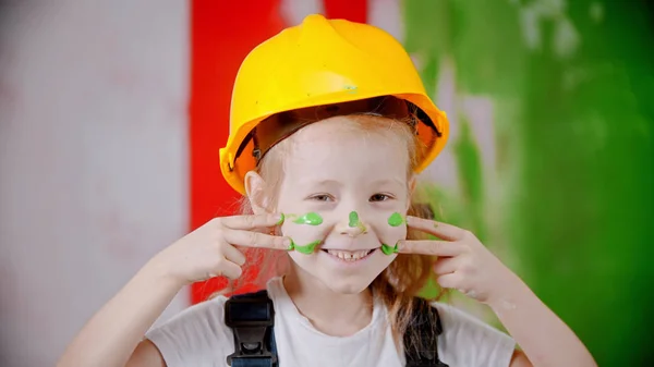 Glimlachend meisje in een helm houdt een verfroller vast en kijkt naar de camera - haar gezicht bedekken met een groene verf — Stockfoto