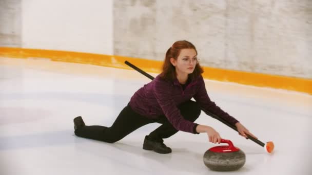 Curling - mladá žena v brýlích tlačí pryč v ledovém poli s žulovým kamenem — Stock video