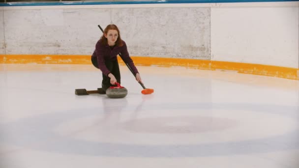 Curling - eine Frau in Brille schiebt sich auf dem Eisfeld mit einem Granitstein in der Hand mit einem Pinsel ab — Stockvideo