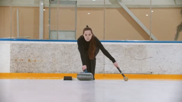 Curling školení - mladá žena s dlouhými vlasy tlačí z tribuny - vede kámen kousnutí s modrou rukojetí — Stock video