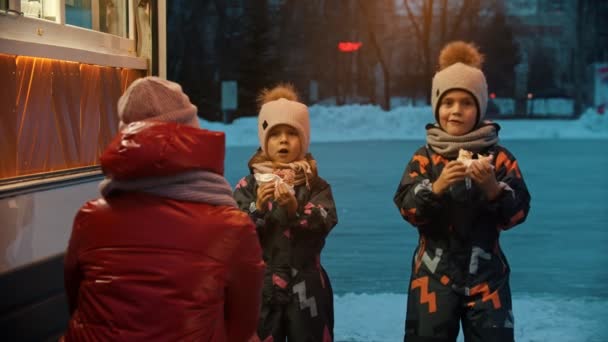 Молодая женщина с детьми пьет горячие напитки и ест пончики рядом с маленьким магазином — стоковое видео