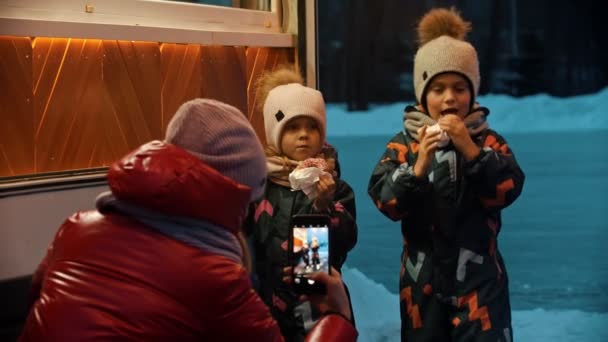 A young woman taking photos of her children drinking hot drinks and eating donuts — Stock Video