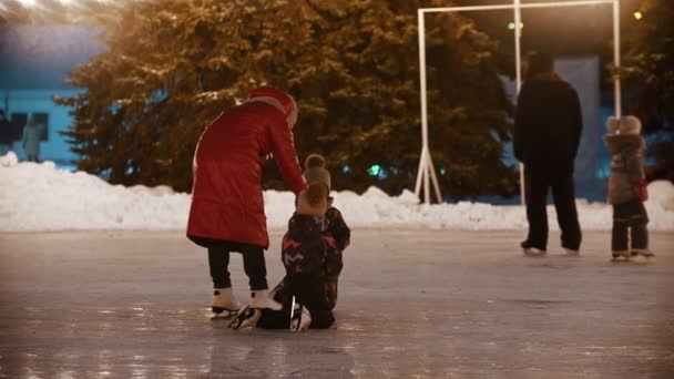 A family of young mother and two kids skating on the decorated ice rink - kids fell on the ice and their mother helping them to get up — Stock Video