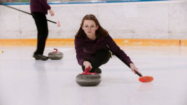 Gözlüklü genç bir kadın buz tarlasına granit bir taşla itiyor.
