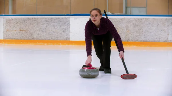Curling treinamento no ringue de gelo - uma jovem mulher de pé na pista segurando uma pedra e escova — Fotografia de Stock