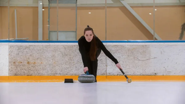 Curling training - una joven con el pelo largo en la pista de hielo sosteniendo una piedra de granito y sosteniendo un cepillo — Foto de Stock