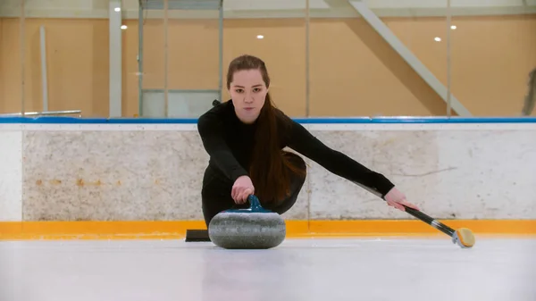 Curling - młoda kobieta z długimi włosami na lodowisku trzymająca granitowy kamień i trzymająca pędzel — Zdjęcie stockowe