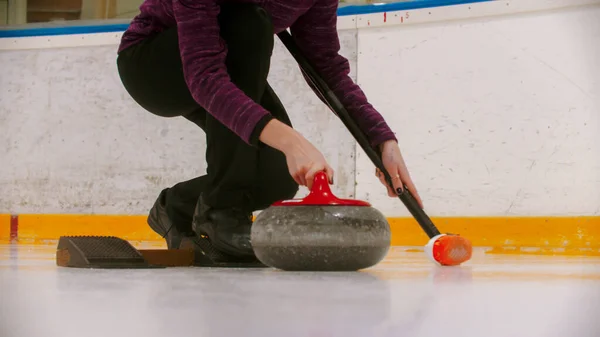 Curling entrenamiento en interiores - una mujer a punto de empujar el soporte en el campo de hielo con piedra de granito —  Fotos de Stock