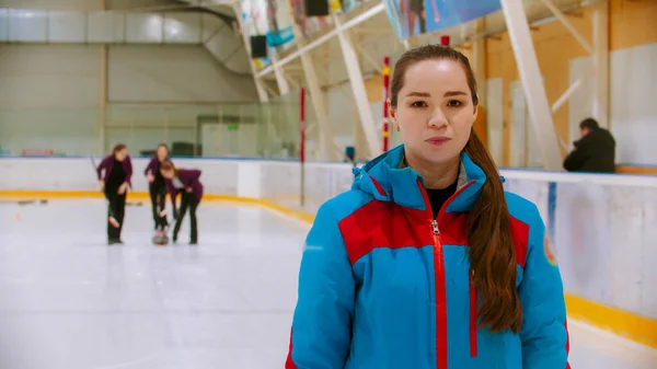 Curling training - the judge in blue jacket standing on the ice rink looking in the camera - her students playing curling on the background