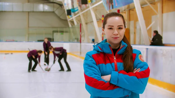 Curling utbildning - leende domare i blå jacka står på ishallen tittar in i kameran - hennes elever spelar curling på bakgrunden — Stockfoto